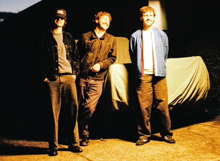 Australian surf rock trio The Grogans standing and smiling outdoors under warm night lighting in a photo by Dylan Jardine.
